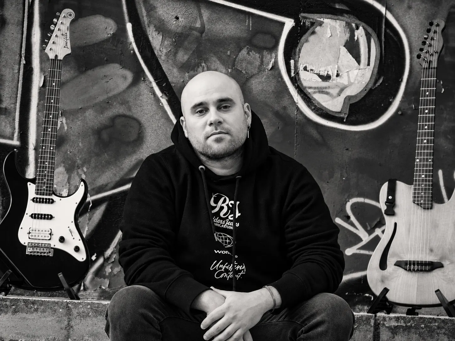 Retrato en blanco y negro de Marquitos Folkis frente a un mural de grafiti, entre una guitarra eléctrica y una criolla